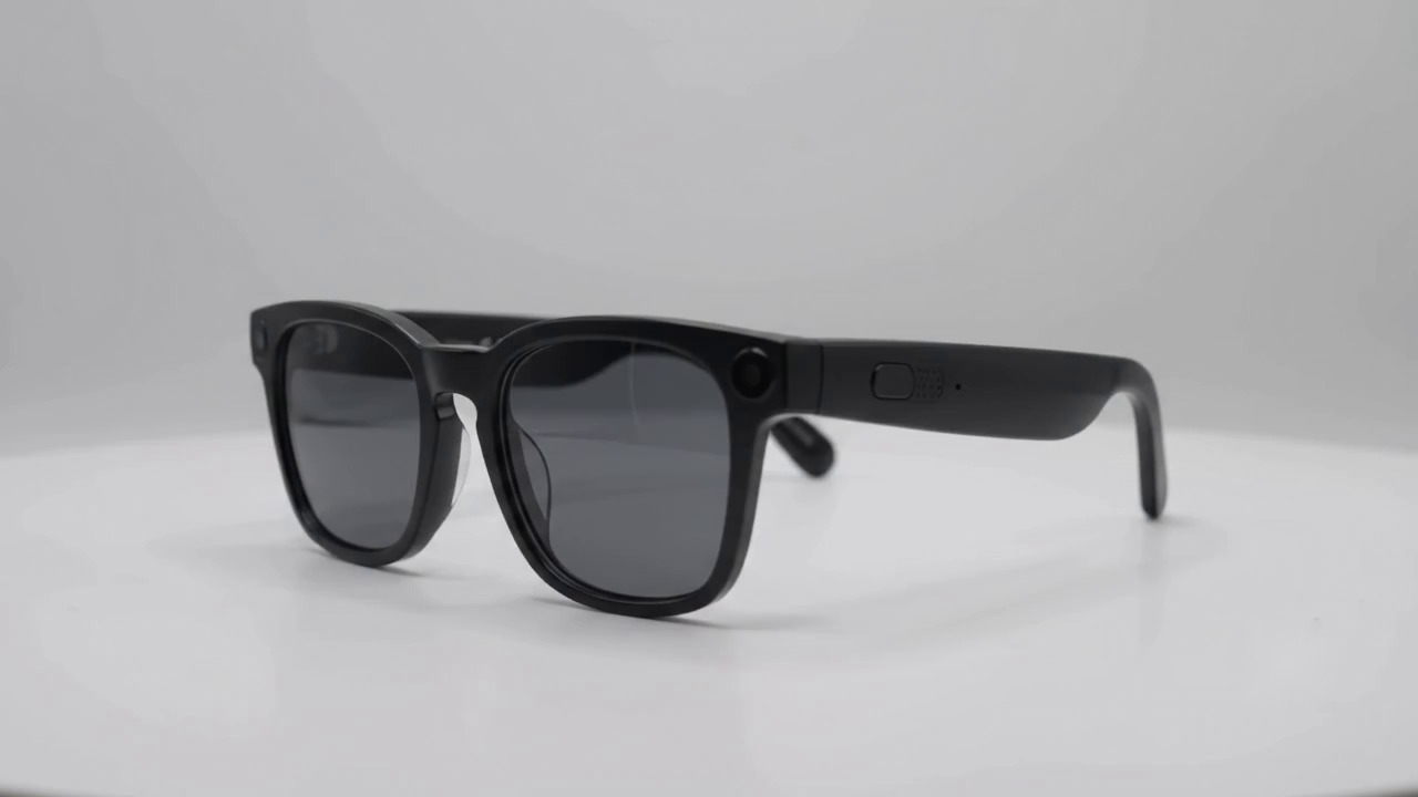 A pair of matte black Blynq glasses in three-quarter view. A recessed button, a speaker grille and a pinhole microphone sit along the temple arm.